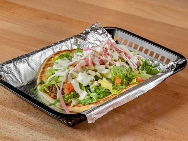 Gyro in a tray topped with lettuce, tomatoes, onions, and white sauce on a wooden surface.