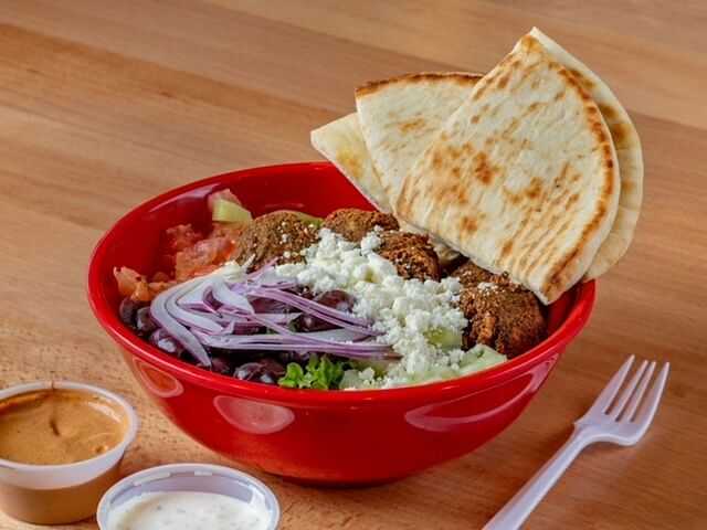 Red bowl with falafel, pita bread, onions, and feta, alongside two dipping sauces and a plastic fork.