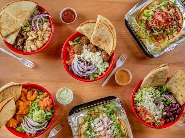 Five bowls of various Mediterranean dishes with pita bread, salads, and sauces on a wooden table.