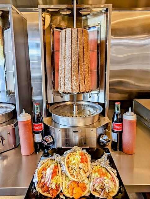 Three shawarma wraps with various fillings, two sodas, and sauces in front of a shawarma rotisserie.